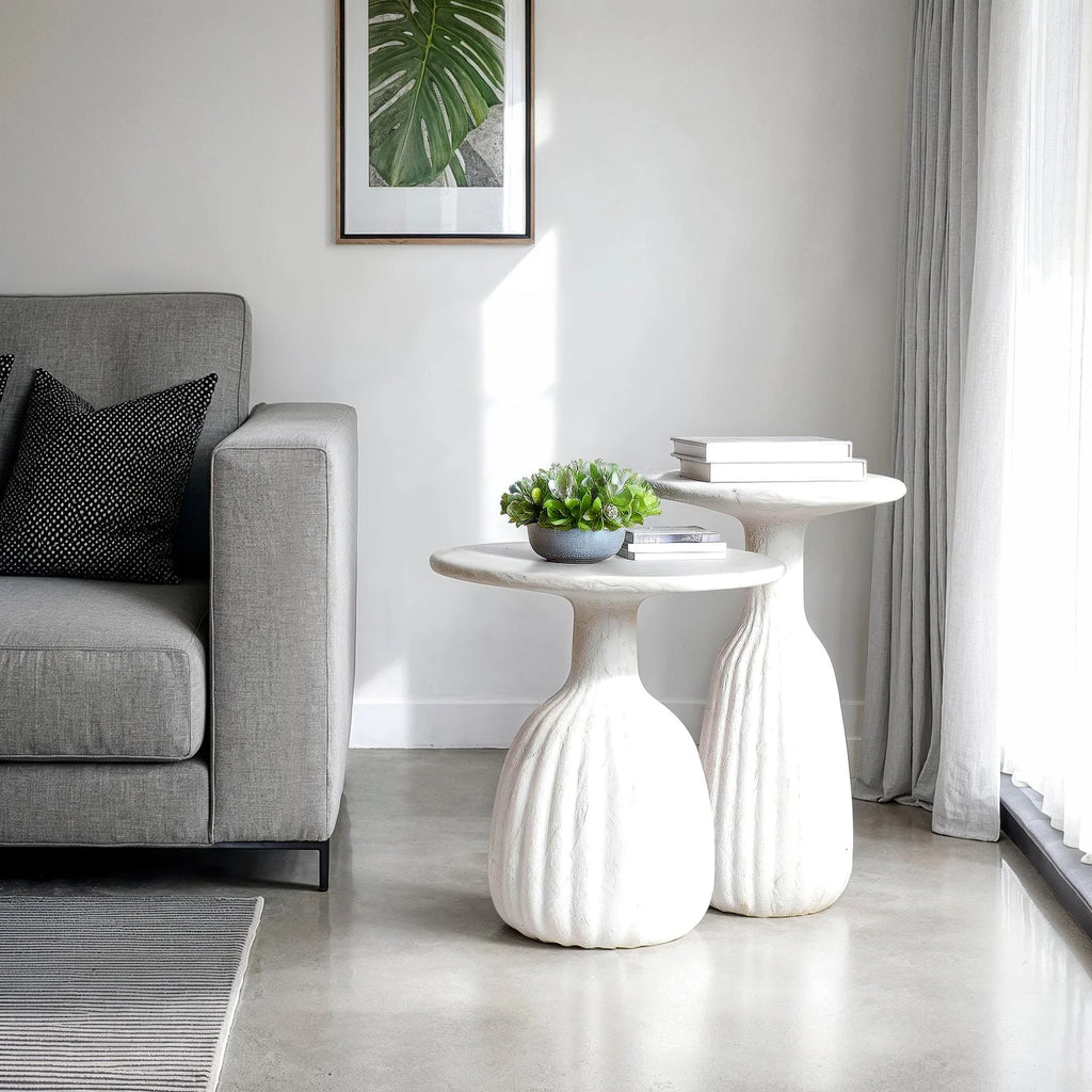 A white accent table with a sculptural base, placed on a floor next to a sofa with a framed picture and a potted plant on top.